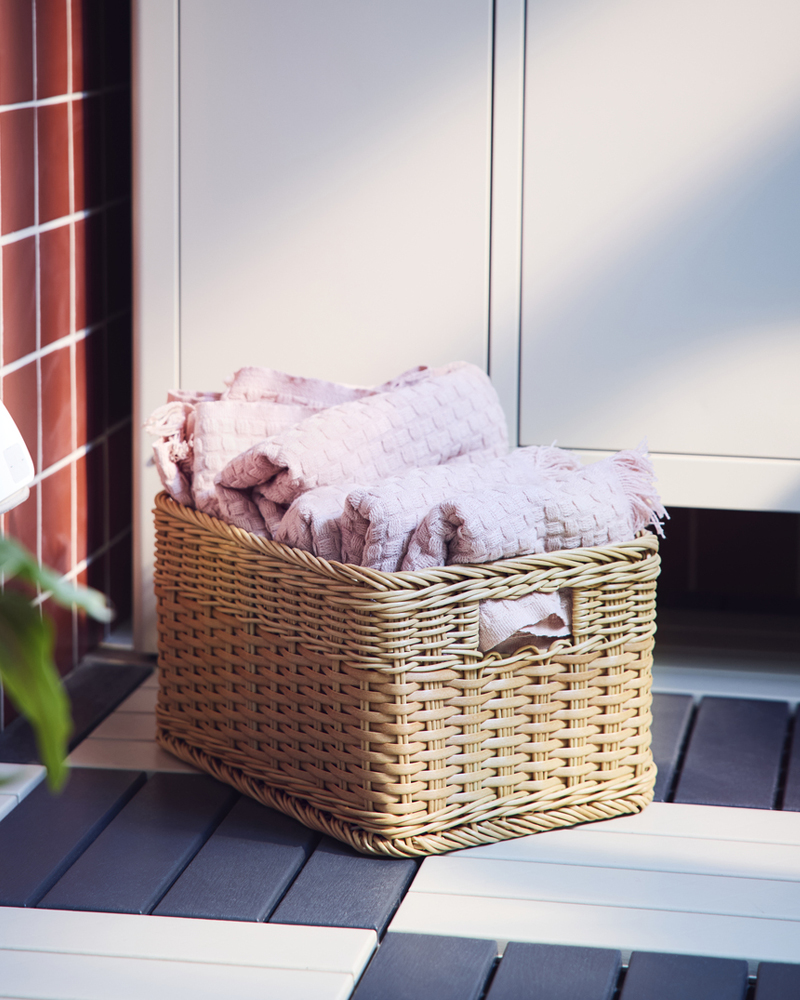A balcony with a natural coloured BEKNA basket made from plastic rattan, filled with cosy throws near a storage cabinet.