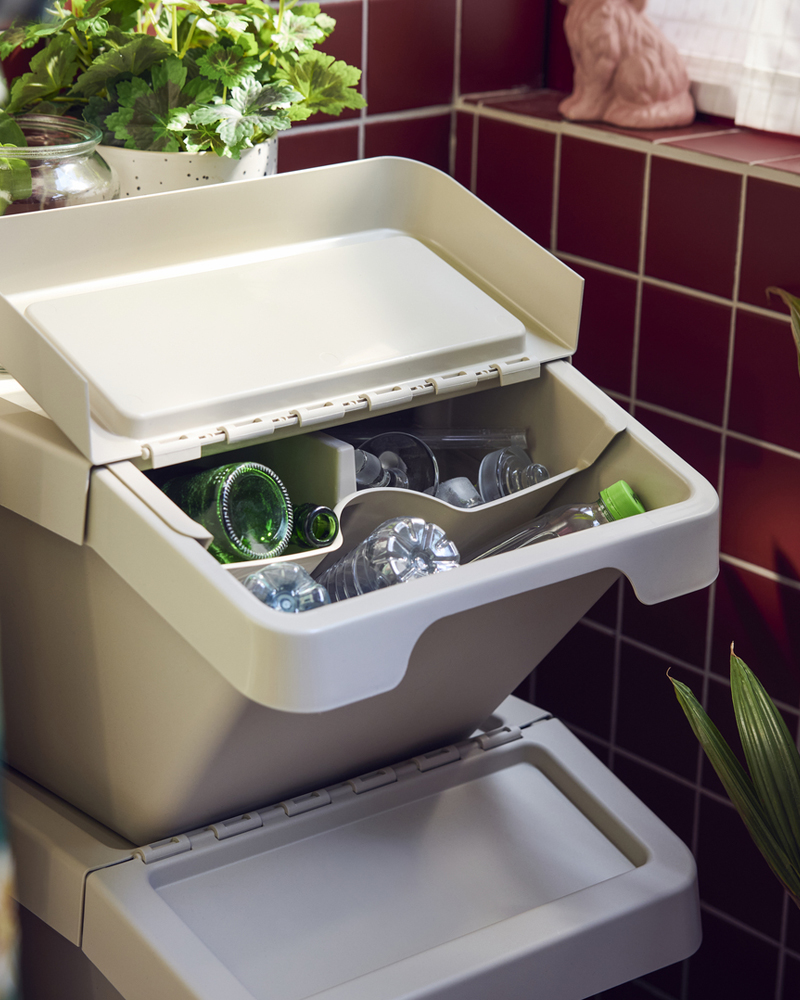 A balcony featuring two beige SORTERA sorting bins with lid, filled with glassware and plastic bottles on a tiled balcony.
