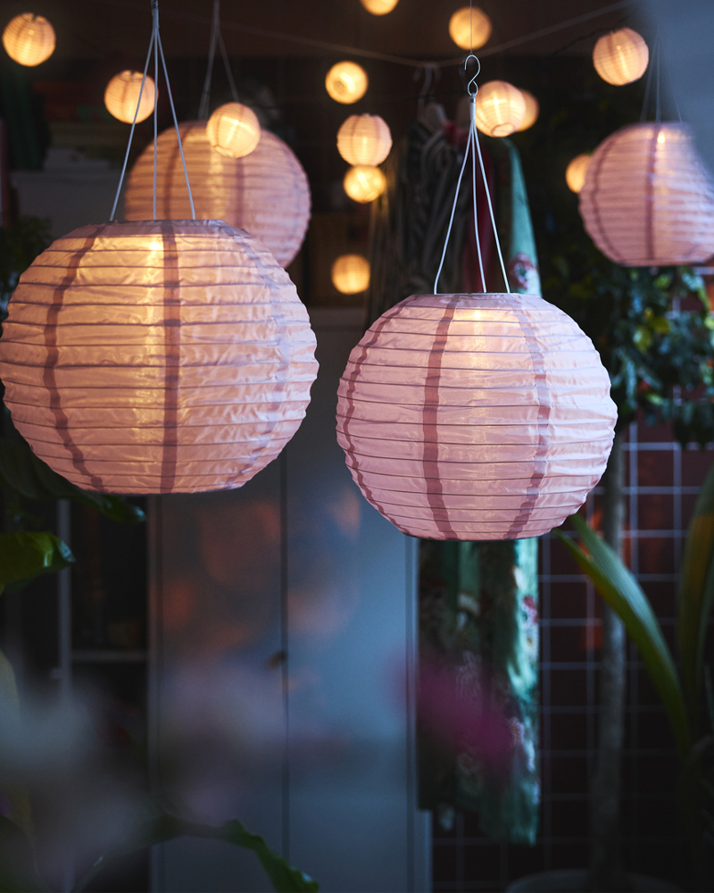 A balcony decorated with lush plants features several pink SOLVINDEN LED solar-powered pendant lamps hanging above.