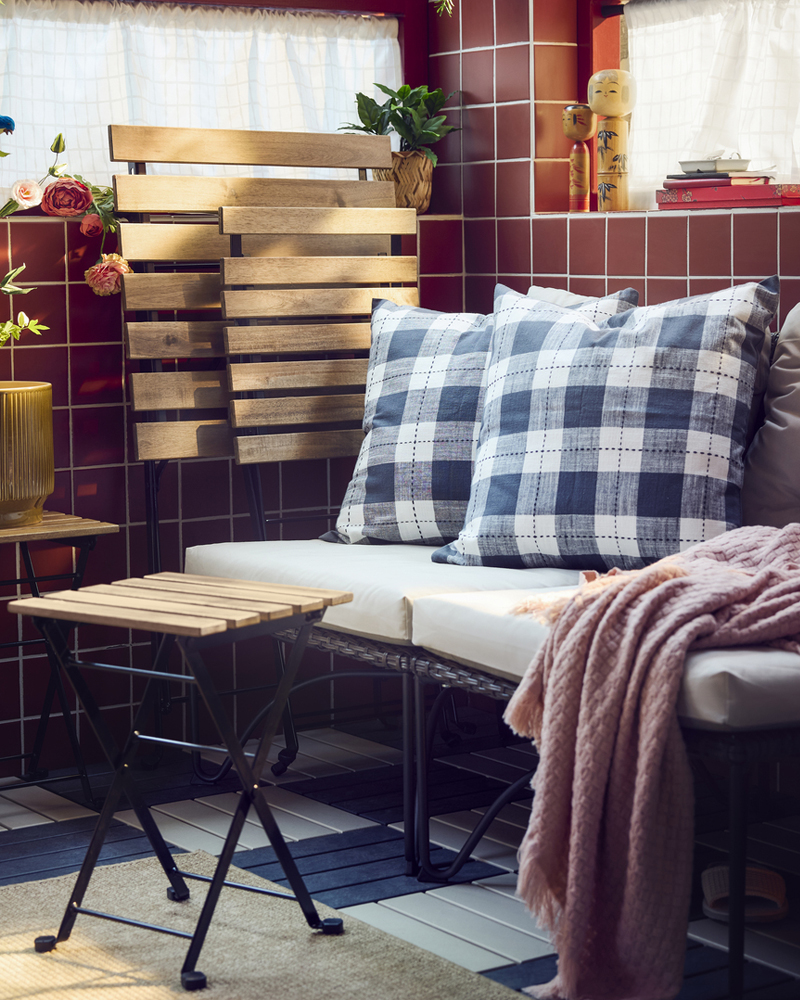A balcony with dark grey JUTHOLMEN easy chair and light grey-beige cushions surrounded by patterned pillows and green plants.