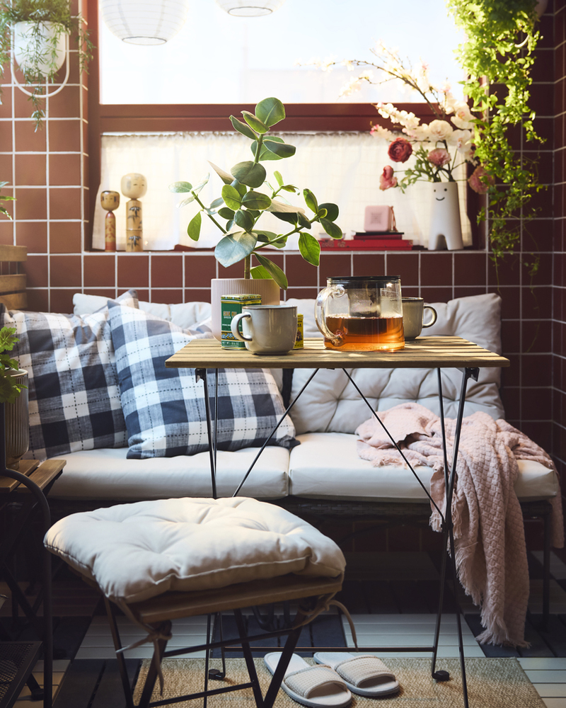 A cosy balcony features a black TÄRNÖ outdoor table, surrounded by plants, cushions, and a glass pot with tea