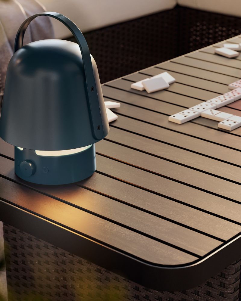 A dark blue VAPPEBY bluetooth speaker lamp for outdoor use on a dark grey VITTSKÄR coffee table. Afternoon light outdoors.
