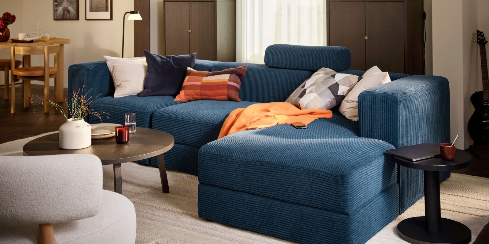 Una sala de estar con un sofá de cuatro plazas JÄTTEBO azul oscuro con chaise longue, complementado con una mesa redonda y elementos decorativos.