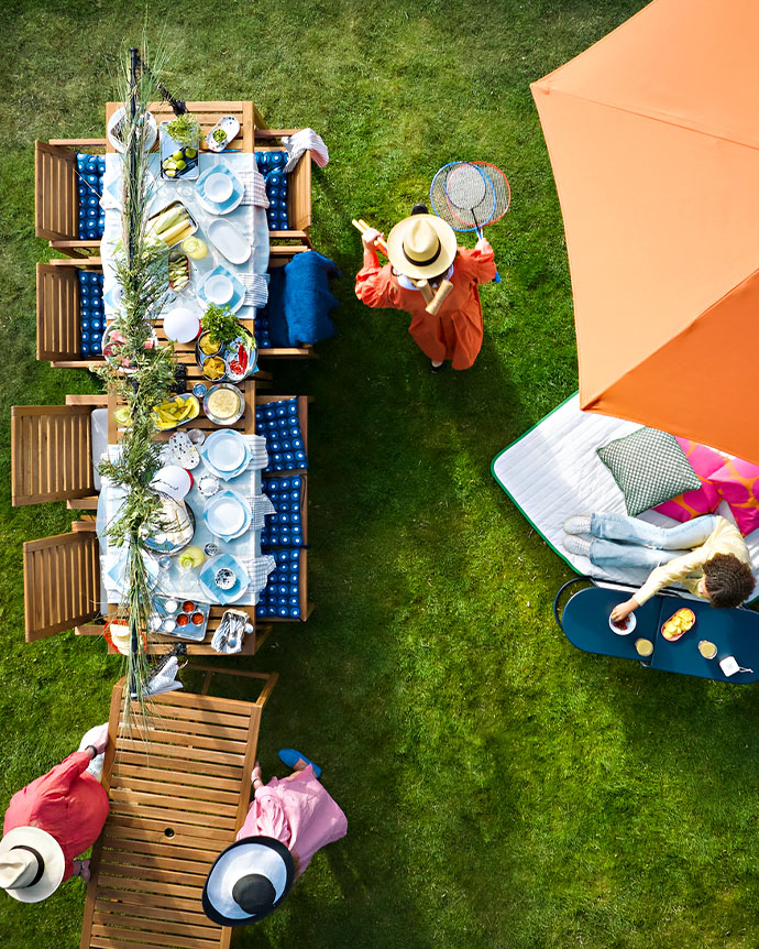 Vista aérea de mesa de madera en jardín con vajilla azul, sombrilla naranja y personas disfrutando al aire libre.