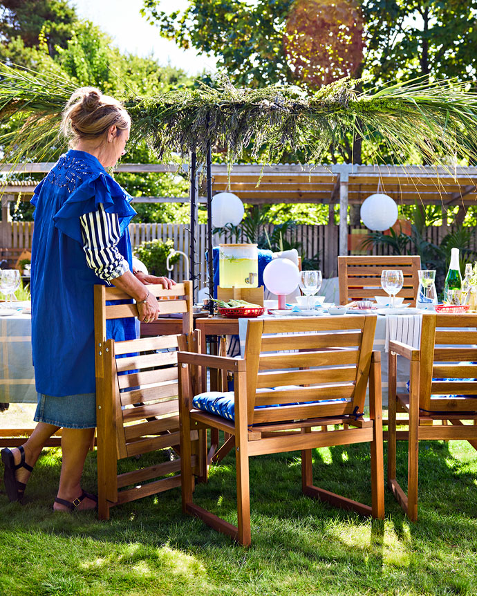 Mujer preparando una mesa de madera con sillas plegables en un jardín soleado, decorado con guirnaldas de vegetación.