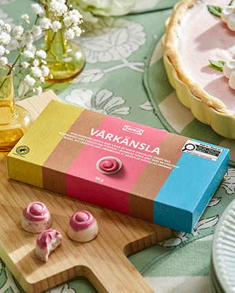 Caja de bombones VÅRKÄNSLA de chocolate rosa y blanco sobre una tabla de madera, rodeada de flores blancas y un mantel floral verde salvia.