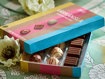 Caja abierta de bombones variados VÅRKÄNSLA que muestra chocolates negros, con leche y blancos con dibujos sobre un mantel floral verde.
