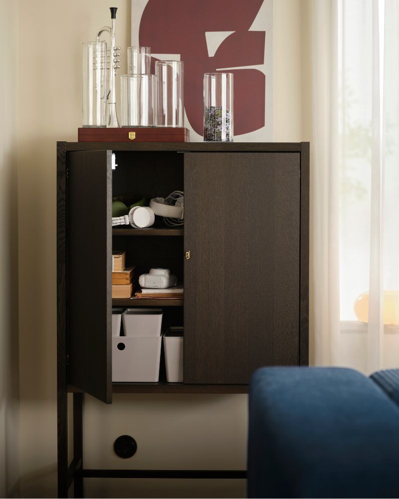Una pared de la sala de estar con un armario STOCKHOLM 2025 de color marrón oscuro que contiene varios accesorios junto a una ventana con cortinas transparentes.