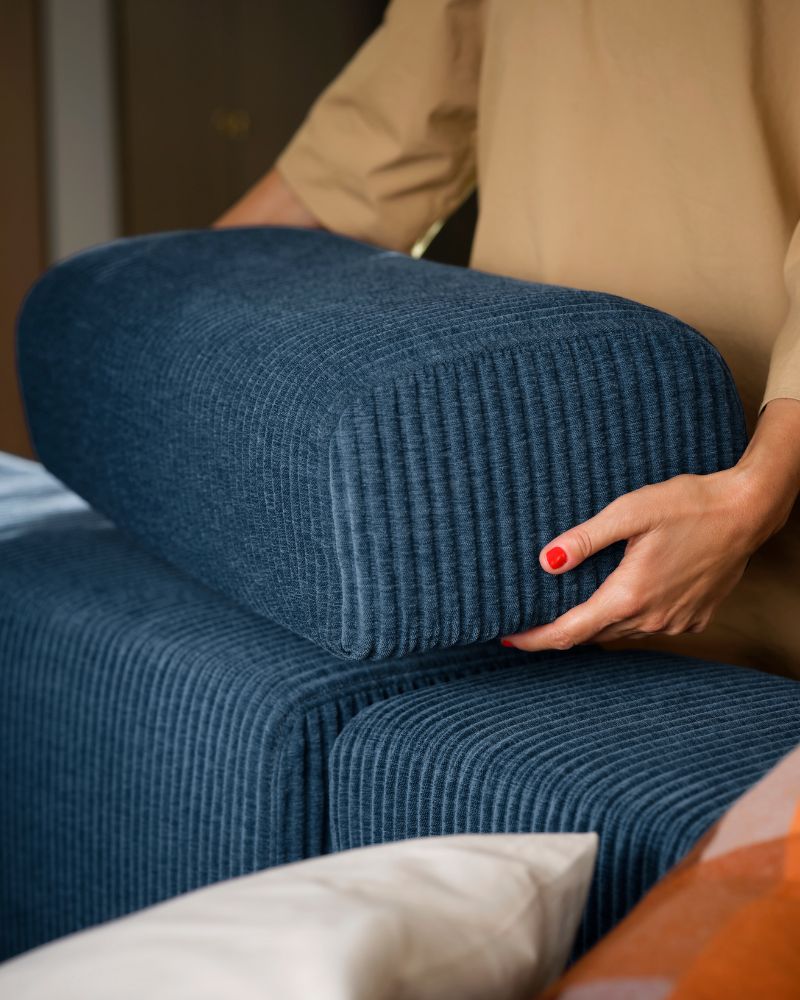 Una persona de pie junto a un sofá modular de cuatro plazas JÄTTEBO azul oscuro coloca un reposacabezas suelto sobre el respaldo.