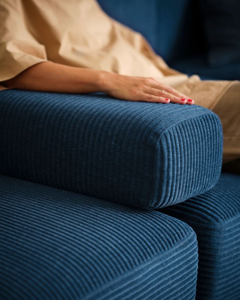 Una persona se sienta en un sofá modular de cuatro plazas JÄTTEBO azul oscuro con el brazo apoyado en un reposabrazos suelto colocado en el sofá.