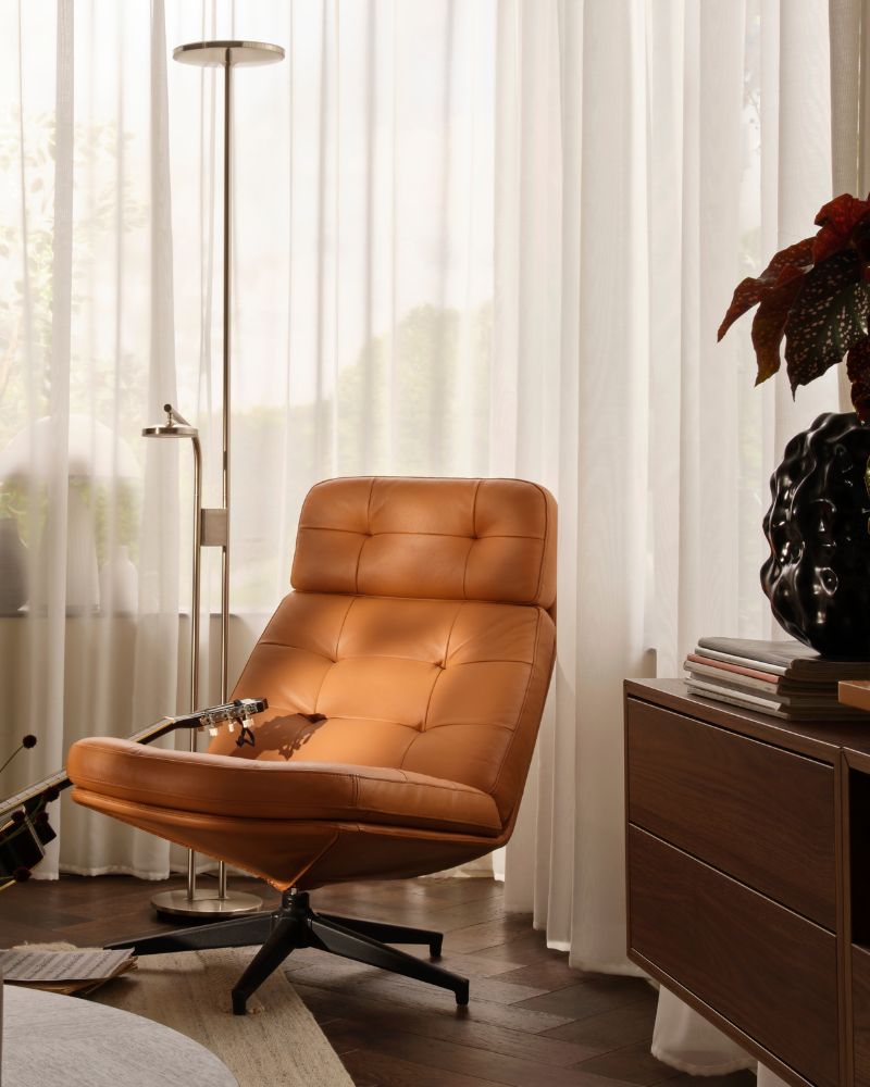 Un rincón del salón con un sillón giratorio HAVBERG de color marrón dorado junto a una gran ventana con cortinas transparentes STENFRÖ.