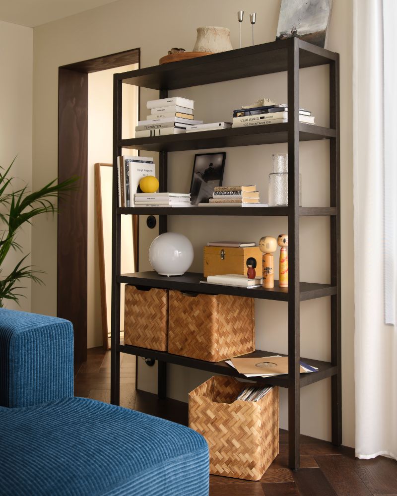 Una sala de estar con una estantería STOCKHOLM 2025 con revestimiento de roble marrón oscuro. Está llena de objetos decorativos, libros y cestas.