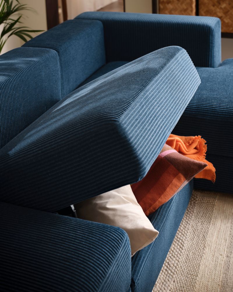 Una sala de estar con un sofá JÄTTEBO de cuatro plazas en azul oscuro, con el asiento central elevado, con un par de cojines y una manta.