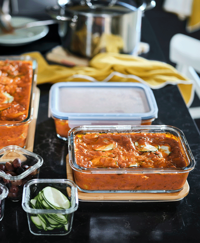 Recipientes de vidrio IKEA 365+ con guiso de verduras y calabacín en rodajas sobre una encimera oscura con un paño amarillo.