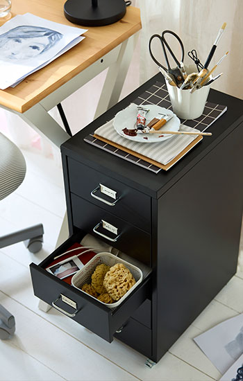 Black HELMER drawer unit on castors beside a BOLLPOJKE desk, holding art supplies with one drawer open