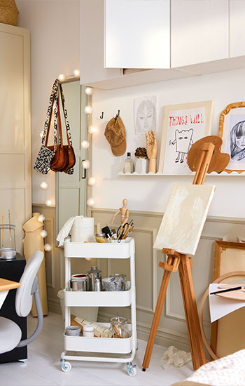 A creative crafting area featuring a white RÅSKOG trolley, holding art supplies next to a desk and an easel in a bright room.
