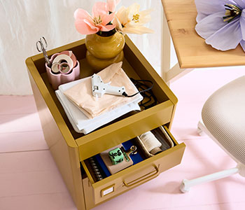 IDÅSEN golden-brown drawer unit on castors with stationery and crafting tools on top, next to a desk with a paper flower
