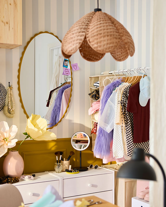 A dressing area with a dark yellow DAGGKAPRIFOL mirror reflecting light, surrounded by clothes on hangers and a flower vase.