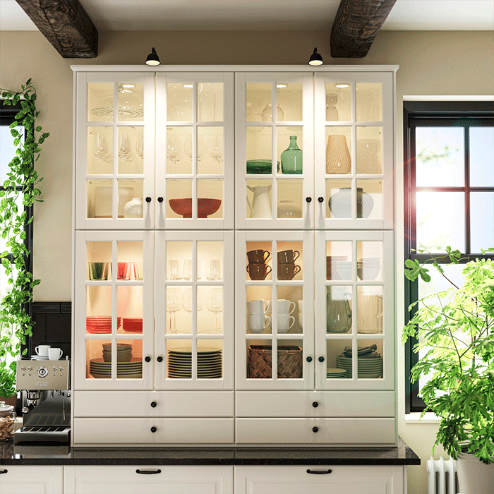 Alacena blanca de puertas de vidrio con iluminación interior, organizando vajilla, copas y tazas entre dos ventanas luminosas.