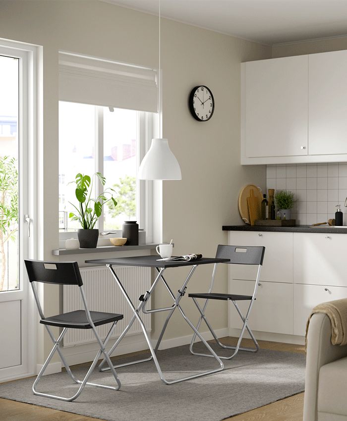 Zona de comedor pequeña en una cocina luminosa con una mesa plegable negra y dos sillas a juego sobre una alfombra gris, situada bajo la ventana.
