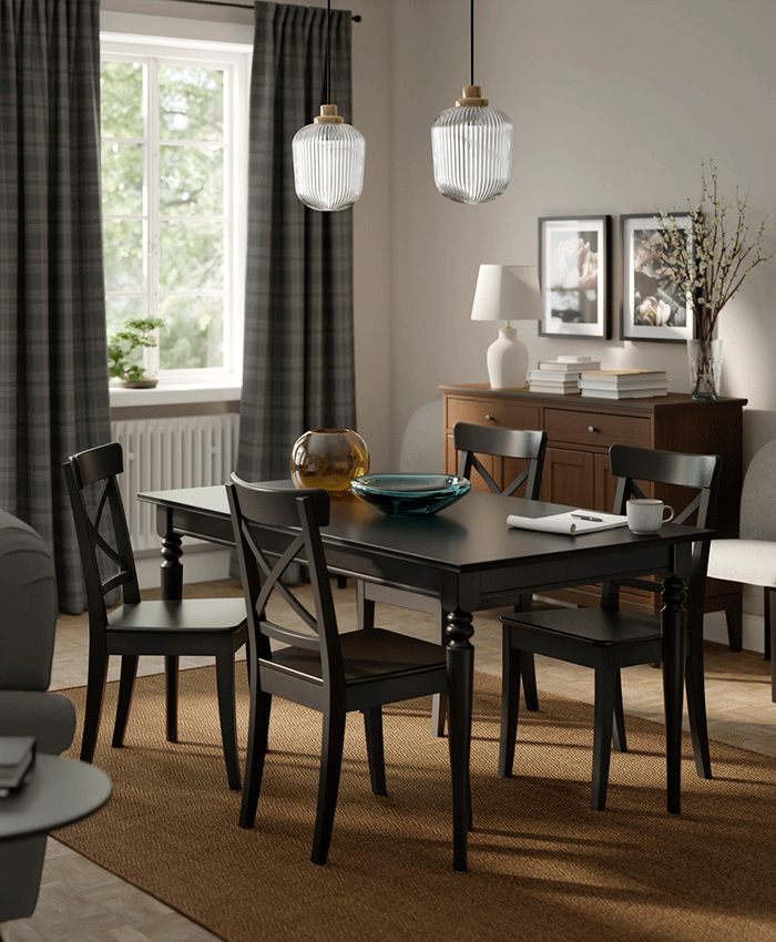 Black wooden dining table with four X-back chairs on a brown rug. A dark wood sideboard and fluted glass pendant lamps create a cozy, moody atmosphere.