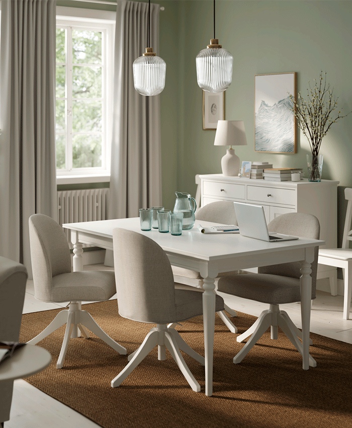 White wooden dining table with four upholstered swivel chairs in beige. Green walls, white sideboard, and light-filtering curtains offer a bright, modern look.