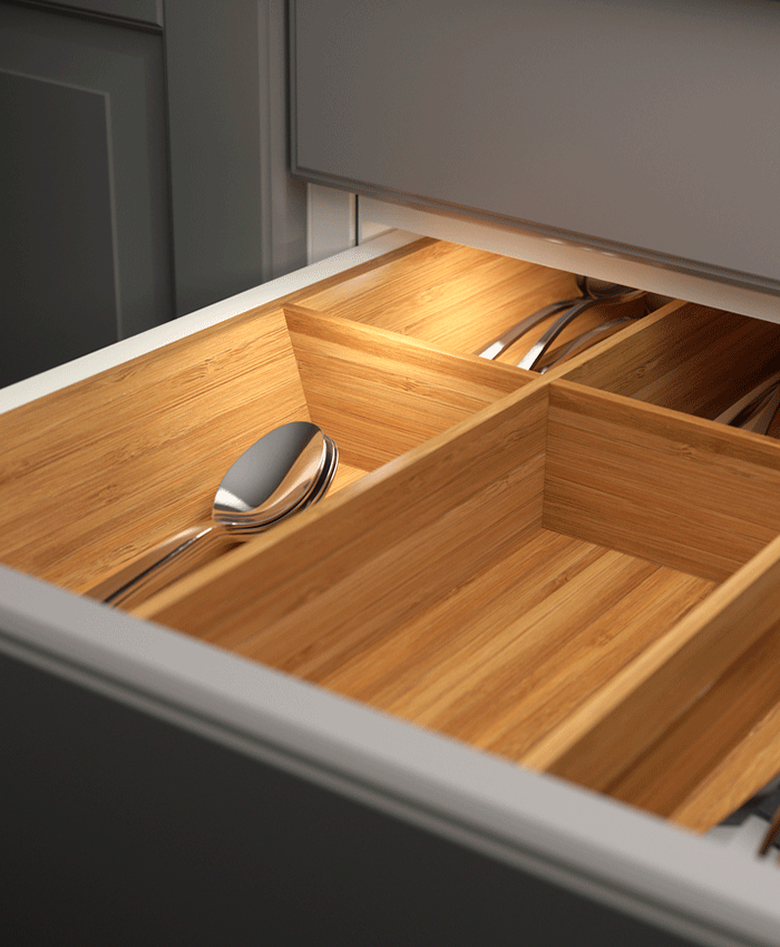 Open kitchen drawer with a bamboo cutlery organizer illuminated by an integrated LED light strip. Silver spoons and forks are neatly arranged in the wooden compartments.