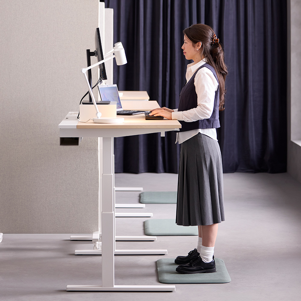 Escena de oficina con una persona de pie utilizando un escritorio MITTZON blanco con chapa de abedul en modo alto, dentro de una configuración de computadora.