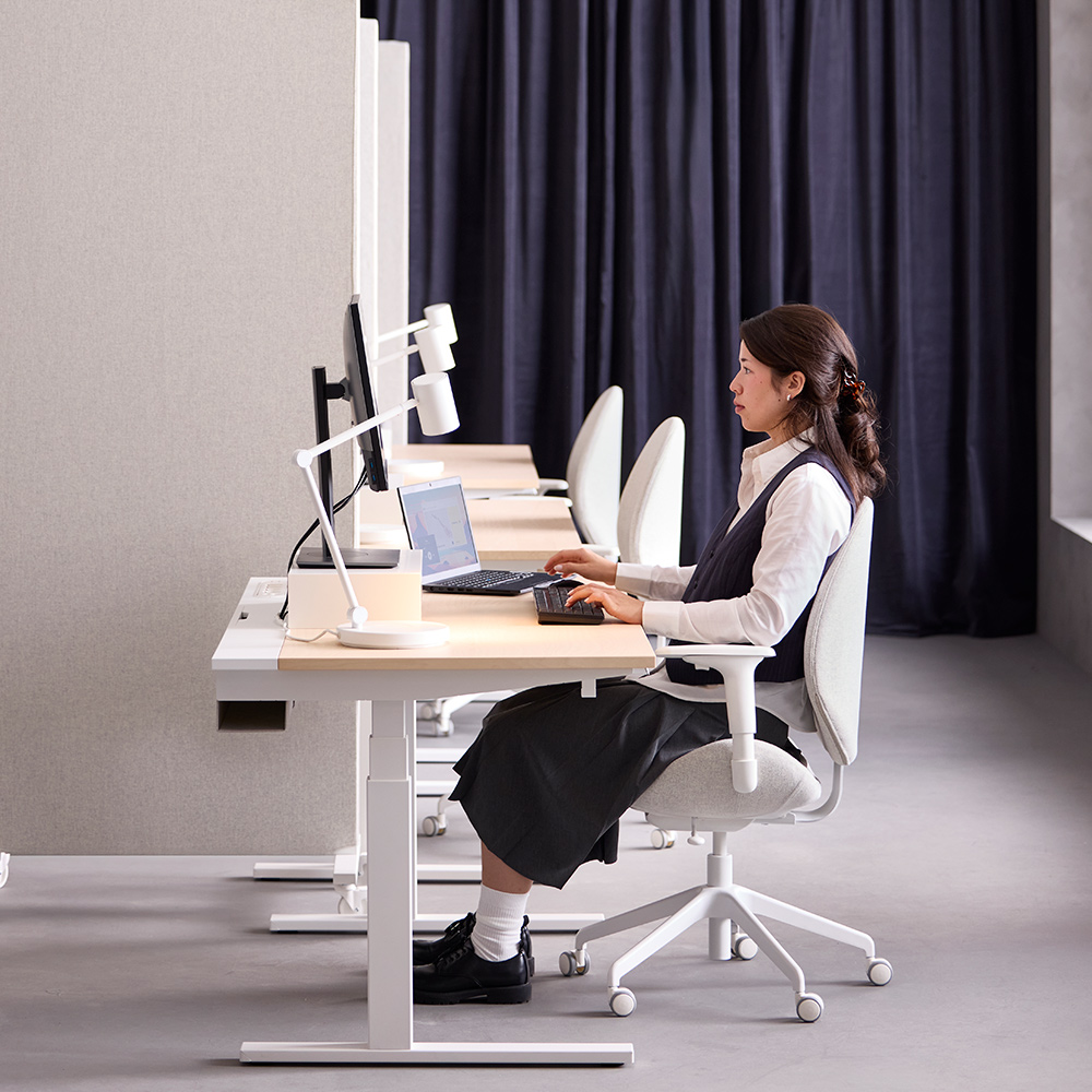 persona utilizando un escritorio MITTZON blanco con chapa de abedul en modo bajo, sentada frente a una configuración de computadora.