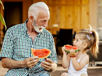 Close-up of an older man and a young girl eating fresh watermelon slices on a sunny terrace with joyful smiles.