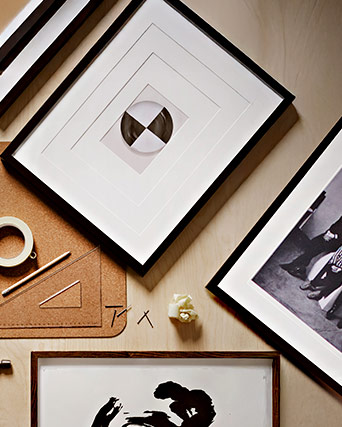 Black picture frames on a light wood table next to measuring tools and a cork base, creating a workspace for home decoration.