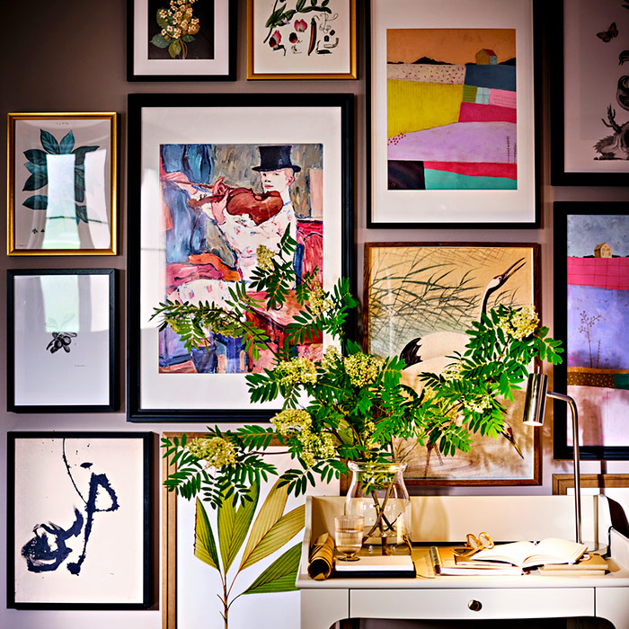 Mauve wall featuring a gallery of diverse framed art above a white desk with a lamp, books, and a glass vase filled with fresh green branches.