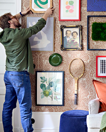 Person hanging frames and vintage items, like a wooden racket, on a wall with classic floral wallpaper, creating a personalized gallery display.