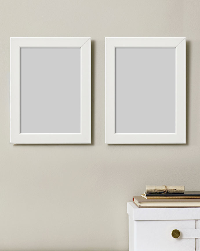 Pair of empty white frames on a beige wall. Below, stacked books and a white storage box with a golden knob.