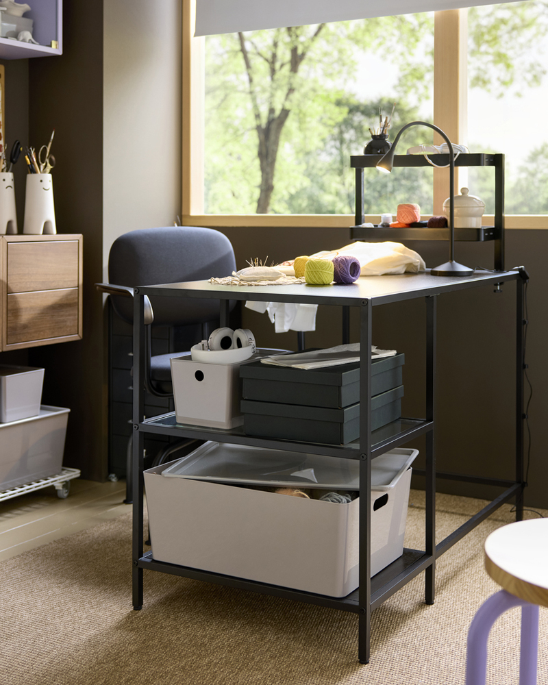 Espacio de trabajo con un escritorio VITTSJÖ marrón oscuro con superficie de cristal, equipado con cajas de almacenamiento y materiales para manualidades.