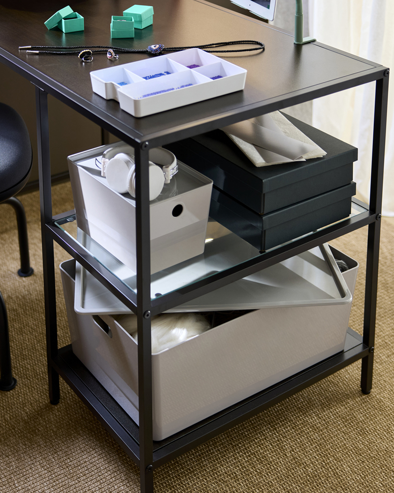 Primer plano de un espacio de trabajo con un escritorio VITTSJÖ marrón oscuro con estantes de cristal, cajas de almacenamiento y materiales para manualidades.