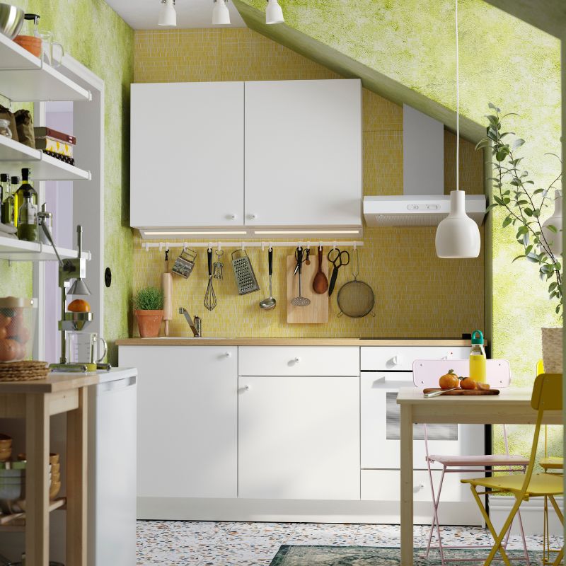 Cocina luminosa y compacta con gabinetes blancos, salpicadero de mosaico amarillo y luz natural. Los utensilios colgantes y las plantas aportan calidez y funcionalidad.