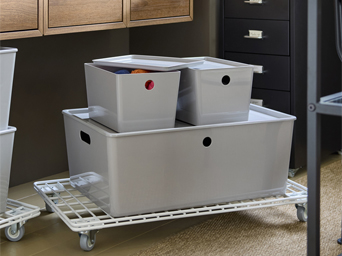 Close-up of light grey KUGGIS storage box under wall cabinets, storing office supplies.