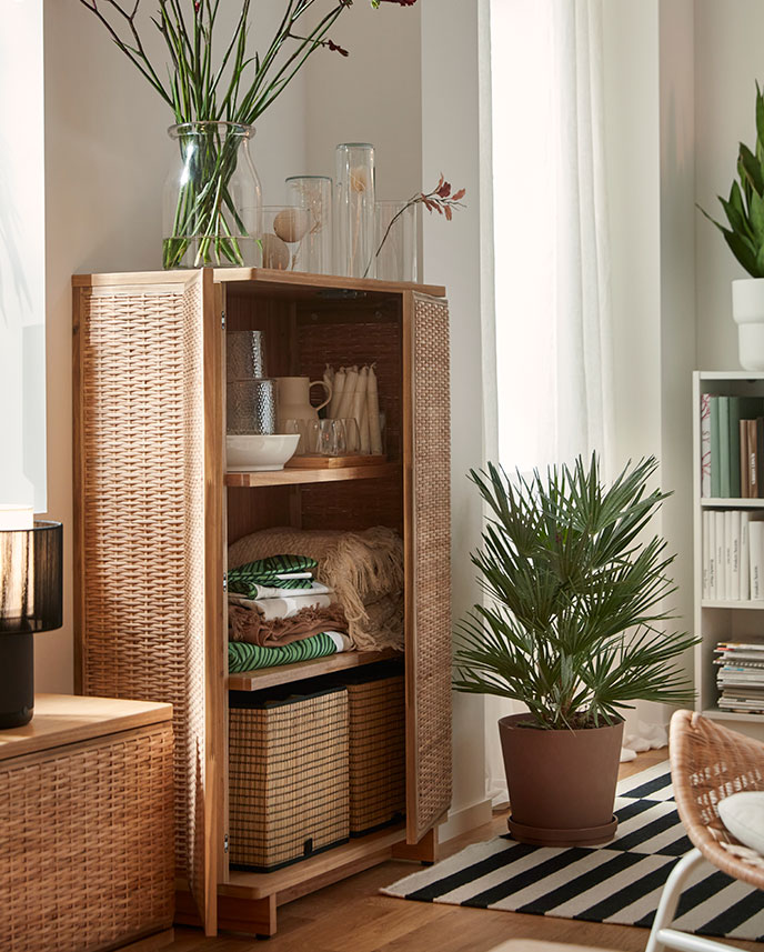 Una luminosa sala de estar con un mueble FRYKSÅS de ratán, en el que se exponen objetos decorativos y que está rodeado de plantas.
