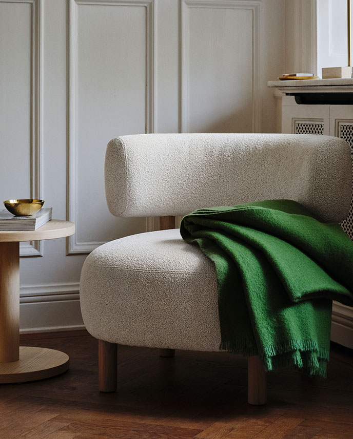 Un rincón luminoso del salón decorado con un sillón STOCKHOLM 2025 gris y blanco, una manta verde y una mesa auxiliar de chapa de pino.