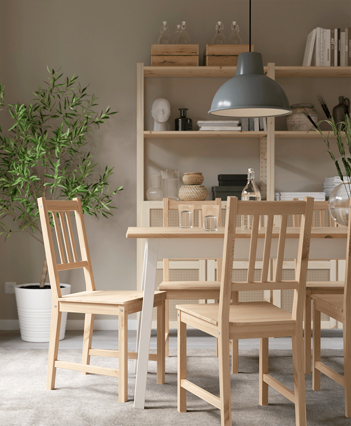 Conjunto de comedor de madera clara con cuatro sillas y mesa a juego en un salón luminoso con una planta grande, estantería y lámpara gris.