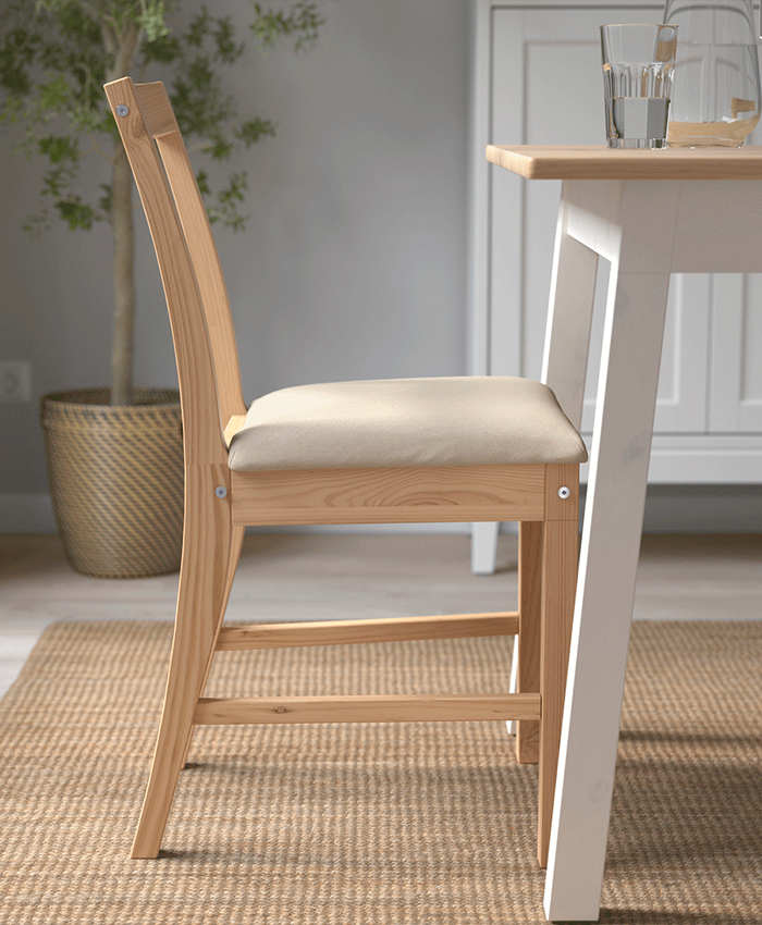Vista lateral de una silla de madera clara con asiento acolchado beige, situada junto a una mesa blanca sobre una alfombra de fibras naturales.