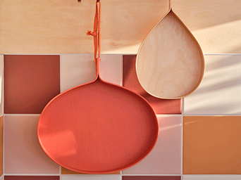 Two teardrop-shaped trays in orange and natural wood hanging on a wall of terracotta, pink, and white square tiles.
