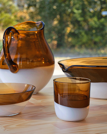 Amber and white mouth-blown glass pitcher, bowl, and tumbler set on a light wood table, featuring a translucent top and opaque base.