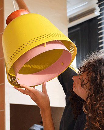 Person adjusting a modern yellow pendant lamp with an inner pink ring and decorative perforations, creating a warm and colorful atmosphere.