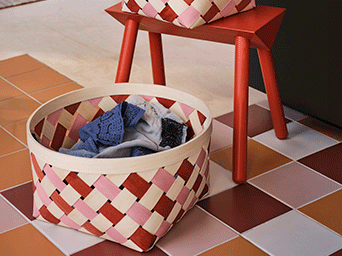 Woven basket with pink, red, and cream strips on a checkered floor, next to a red wooden stool holding another matching basket.