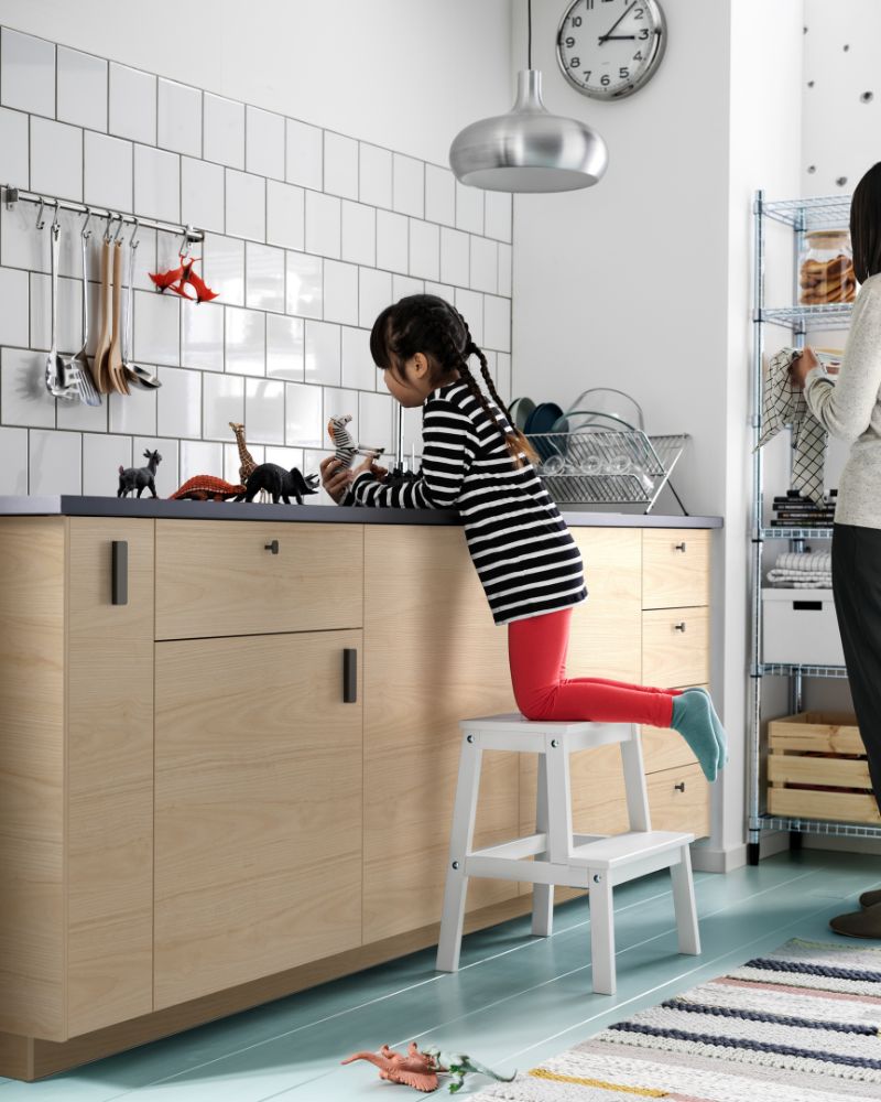 Girl plays with figures in a light wood cabinet kitchen with white tiles, standing on a white step stool.