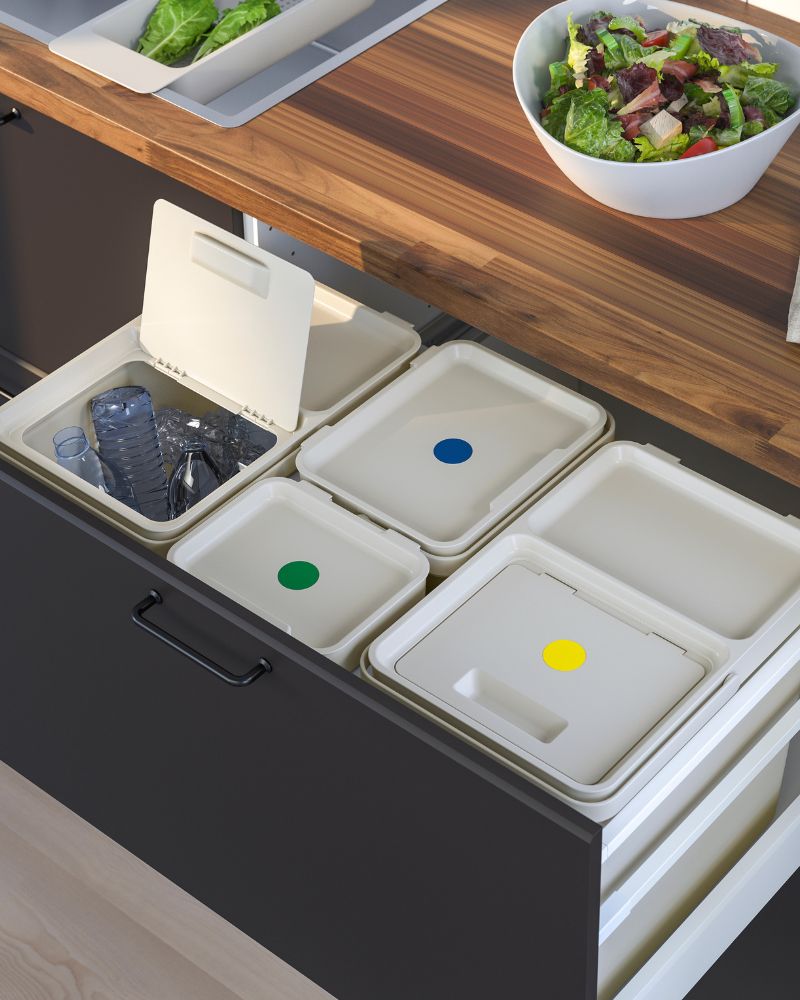 Dark kitchen drawer with white and colorful recycling bins, under a wooden countertop.