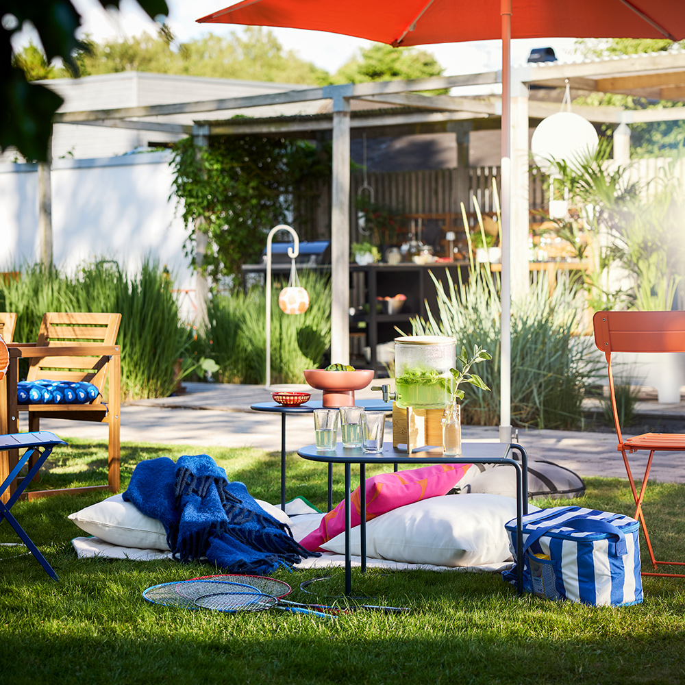 A garden setting with greenery and a sun parasol features a black-blue GRYTSHOLM nest of tables, perfect for outdoor use.