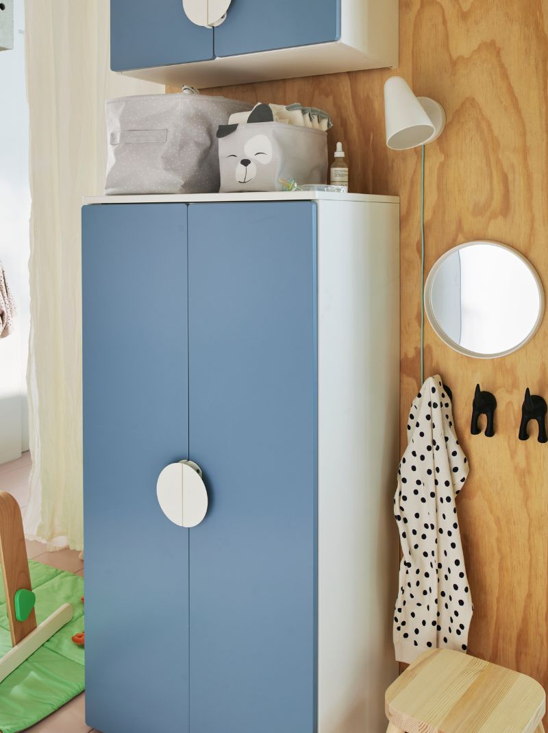 A blue SMÅSTAD wardrobe against a wooden wall with two storage boxes holding baby things on top and a wall light beside 